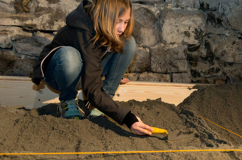 Zoom: Familienausflug Schloss Werdenberg. Kleine ArchäologInnen heben unter der Leitung einer Fachperson entdeckte Fundstücke korrekt aus der Erde, ordnen sie und besprechen deren Bedeutung.