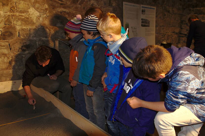 Zoom: Familienausflug Schloss Werdenberg. Kleine ArchäologInnen heben unter der Leitung einer Fachperson entdeckte Fundstücke korrekt aus der Erde, ordnen sie und besprechen deren Bedeutung.
